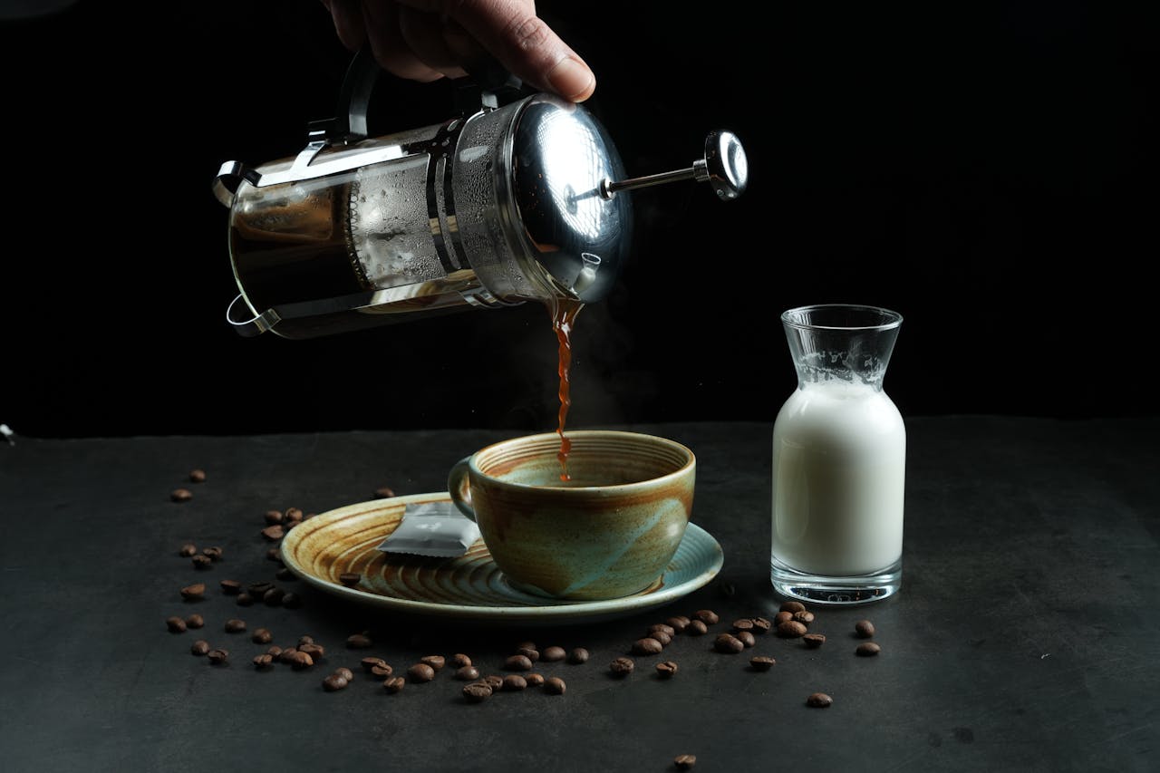 Steaming coffee pouring into a rustic cup with milk jug and scattered beans on a dark surface.